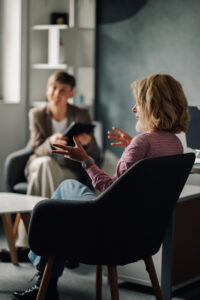 Woman sitting comfortably in an armchair, engaging in conversation with a therapist who is taking notes on a digital tablet during a therapy session